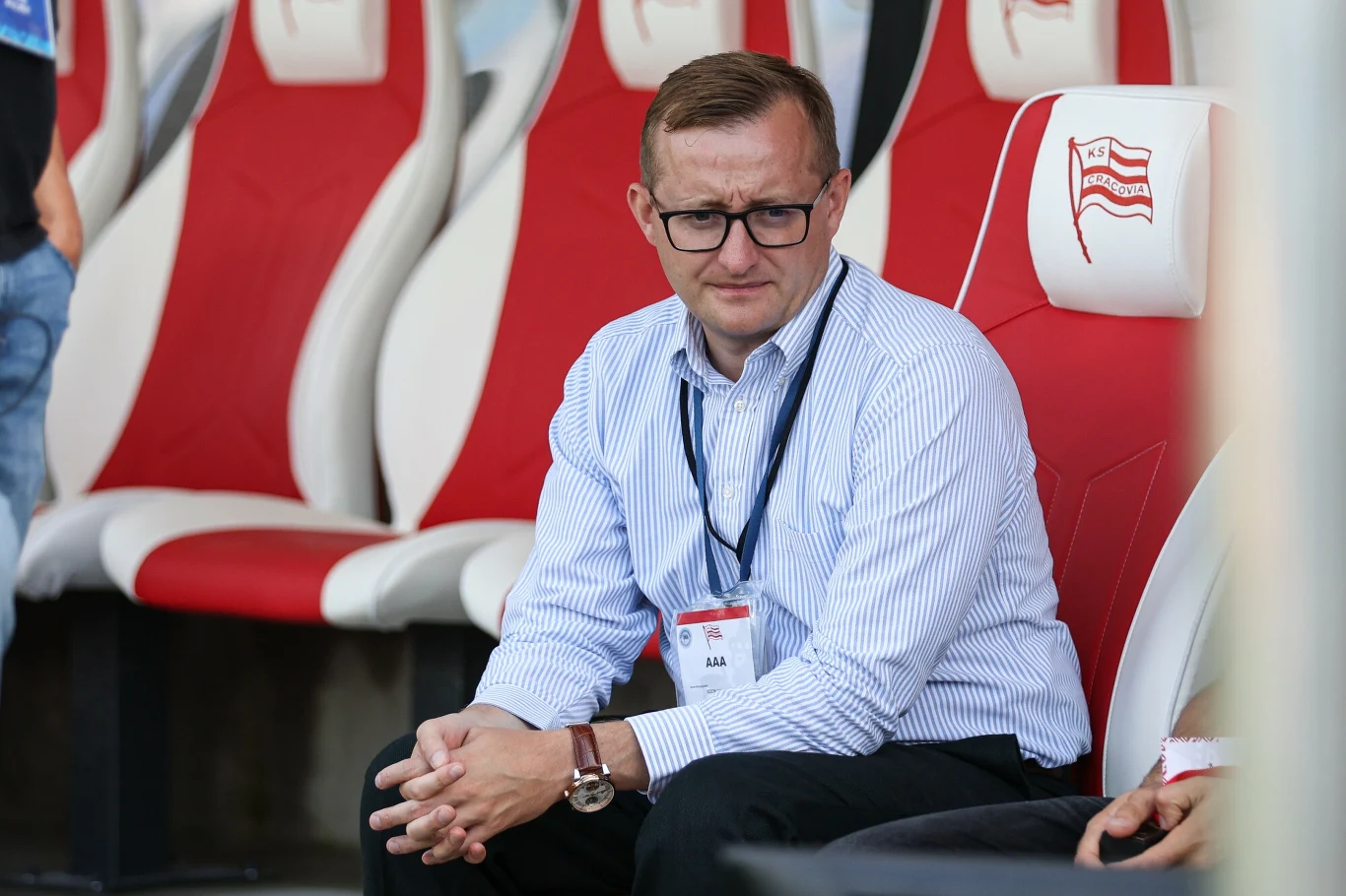 Mateusz Dróżdż, prezes Cracovii Mężczyzna w okularach i eleganckiej koszuli siedzi na stadionowej ławce rezerwowych, obok widać czerwono-białe siedzenia z herbem sportowego klubu.