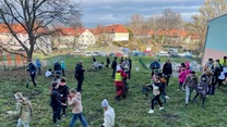 Grupa dzieci i dorosłych uczestniczy we wspólnej aktywności na trawiastym terenie obok budynku, część osób kopie dziury lub sadzi rośliny, widoczne są jesienne drzewa oraz osiedle domków w tle