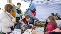 Grupa młodych osób zgromadzona przy stoliku z materiałami edukacyjnymi, notesami, torbami i gadżetami ekologicznymi, uczestnicy są zainteresowani prezentowanymi produktami, atmosfera jest skupiona i pozytywna.