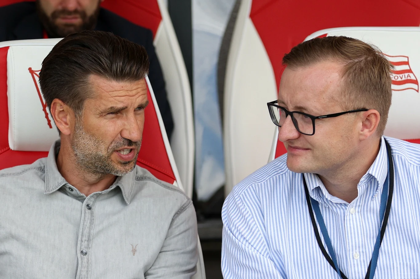 Luka Elsner i Mateusz Dróżdż Dwóch mężczyzn siedzi obok siebie na stadionowych fotelach, jeden z nich mówi, drugi słucha z lekkim uśmiechem, mają na sobie koszule, jeden z nich posiada identyfikator.