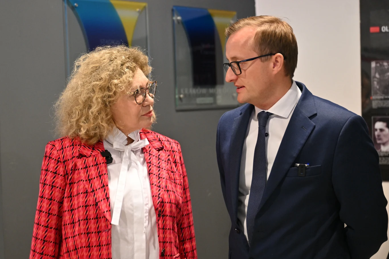 Elżbieta Filipiak i Mateusz Dróżdż Dwoje ludzi prowadzi rozmowę, kobieta w czerwonej marynarce i białej bluzce oraz mężczyzna w granatowym garniturze, oboje w okularach, znajdują się w jasnym wnętrzu z dekoracjami na ścianie.