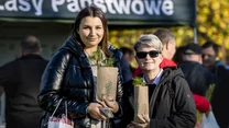 Dwie kobiety ubrane w kurtki trzymają młode sadzonki drzew w papierowych torebkach, stojąc na tle ludzi i stoiska z napisem 'Lasy Państwowe'.