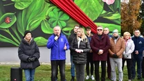 Grupa osób stojąca przed dużym muralem z motywami roślinnymi i wstęgą, jeden z mężczyzn przemawia do mikrofonu, reszta osób słucha. W tle widoczny fragment zieleni i jesienne drzewa.