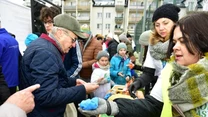 Grupa osób, głównie seniorzy i dzieci, odbierają przedmioty od wolontariuszy przy stoliku na świeżym powietrzu, w tle widać budynki mieszkalne, a atmosfera jest przyjazna i zorganizowana.