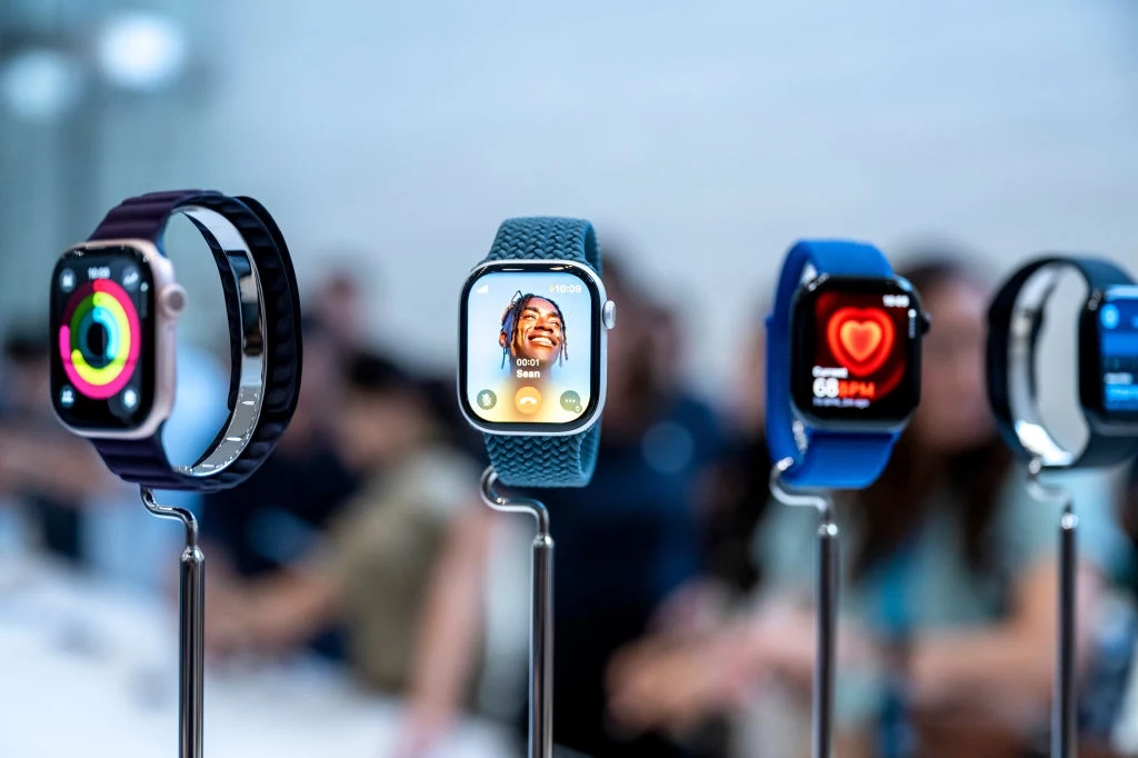 Apple twierdzi, że ich smartwatche są neutralne klimatycznie, bo gigant płaci za sadzenie eukaliptusów w Paragwaju Rząd różnokolorowych smartwatchy prezentowanych na metalowych stojakach, na jednym z nich widoczne kolorowe kółka aktywności, na innym ekran z wizerunkiem uśmiechniętej osoby, tło rozmyte z sylwetkami ludzi.
