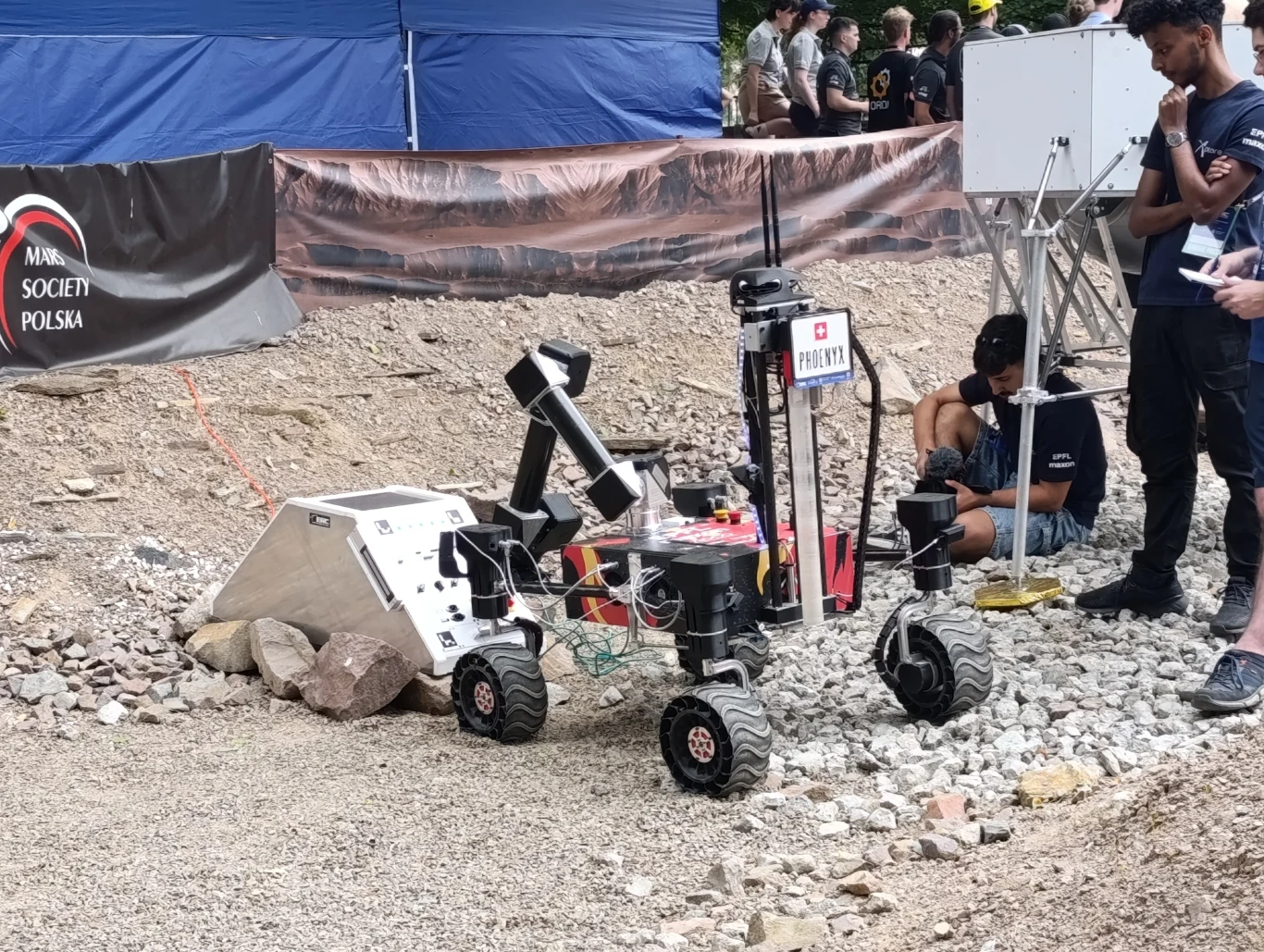 Łazik Phoenyx szwajcarskiego zespołu EPFL Xplore Dwukołowy łazik mobilny z chwytakiem podczas testów na żwirowym terenie, wokół kilku osób obserwujących jego pracę, w tle namiot i baner stowarzyszenia naukowego.