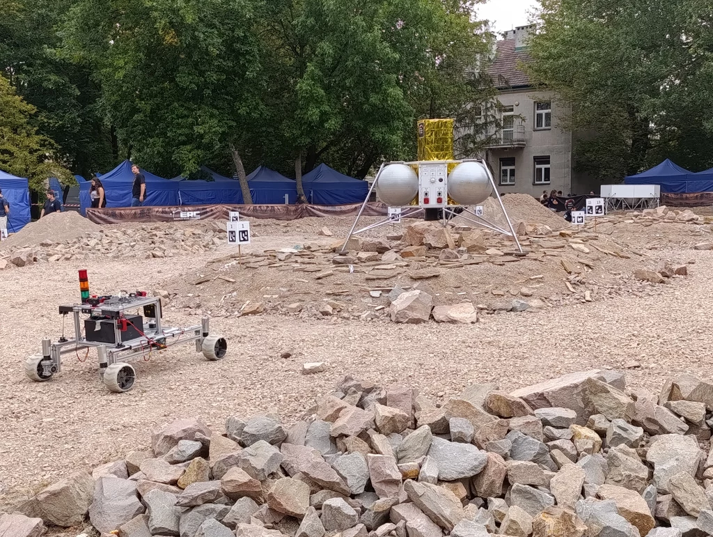 Marsyard, na którym łaziki prezentowały swoje możliwości Robot eksploracyjny porusza się po kamienistym terenie, naprzeciwko lądownika kosmicznego ustawionego w centrum poligonu, wokół rozstawione są namioty oraz niewielka grupa obserwatorów.