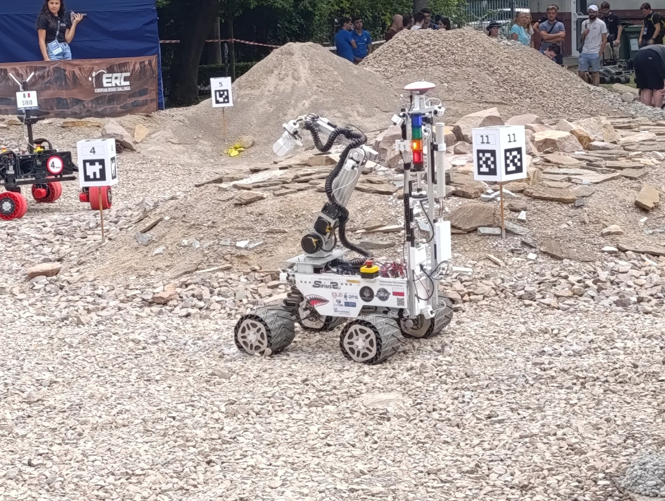 Łazik Sirius II polskiej drużyny z Politechniki Warszawskiej SKA Robotics Robot mobilny z ramieniem manipulacyjnym jadący po kamienistym terenie, wokół widoczne pagórki ziemi, tabliczki z numerami oraz niewielka grupa obserwatorów w tle.