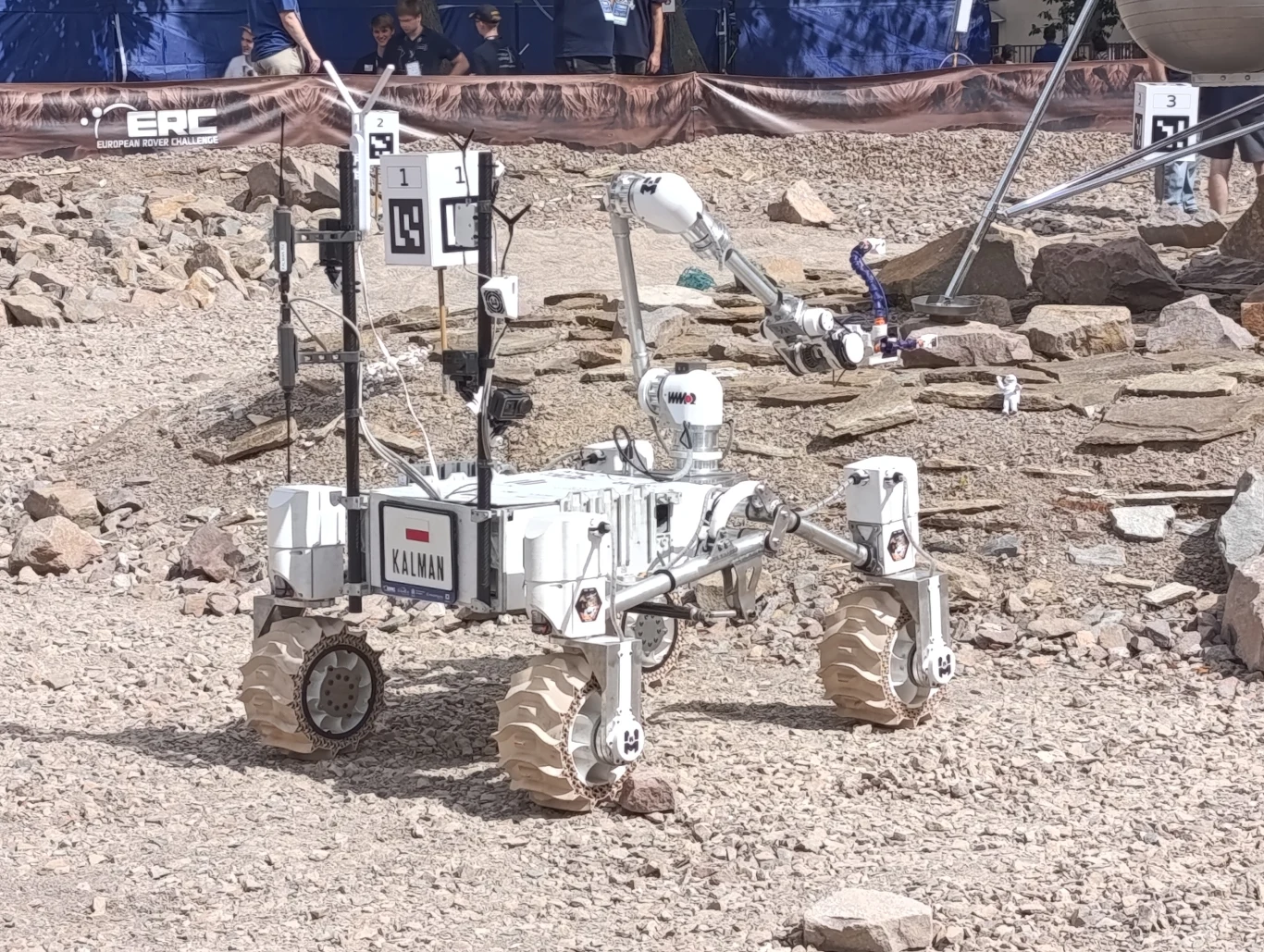 Łazik Kalman polskiej drużyny AGH Space Systems Robot mobilny z manipulatorem poruszający się po kamienistym, pustynnym terenie testowym, wokół widoczne są elementy infrastruktury technicznej oraz bariera z logotypem ERC, w tle kilku obserwatorów.