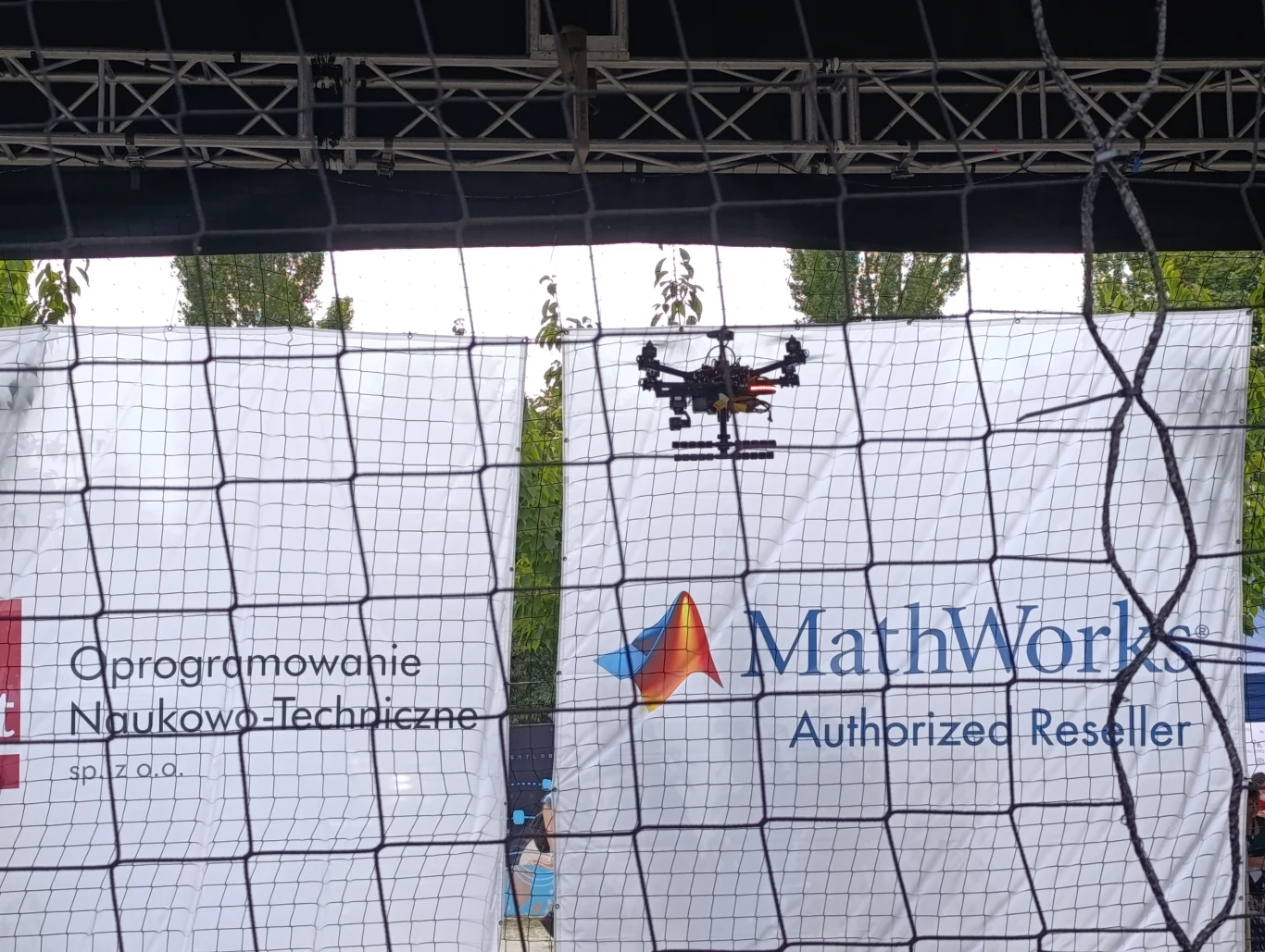 W specjalnie zaprojektowanej klatce drużyny manerwowały swoimi dronami Dron unoszący się w powietrzu za siatką ochronną, w tle banery reklamowe firm związanych z oprogramowaniem naukowo-technicznym oraz sprzedażą oprogramowania matematycznego.