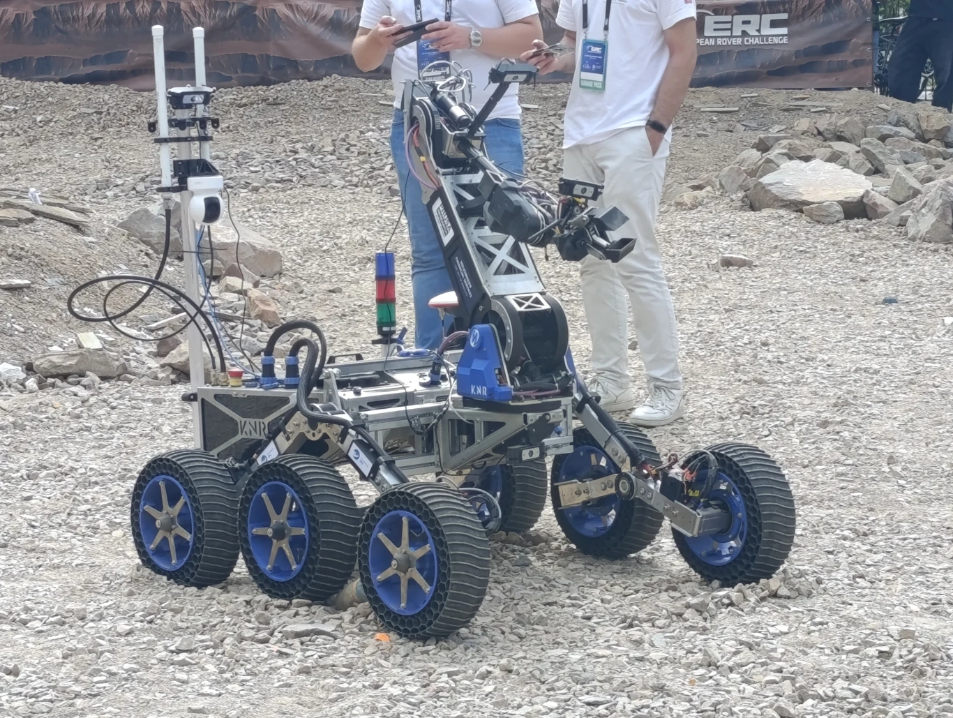 Łazik HAL-062 polskiej drużyny KNR Rover Team z Politechniki Warszawskiej Sześciokołowy łazik robotyczny z licznymi czujnikami i manipulatorami prezentowany na kamienistym terenie przez osoby ubranie w białe koszulki i spodnie, prawdopodobnie podczas konkursu naukowego lub wydarzenia technologicznego.
