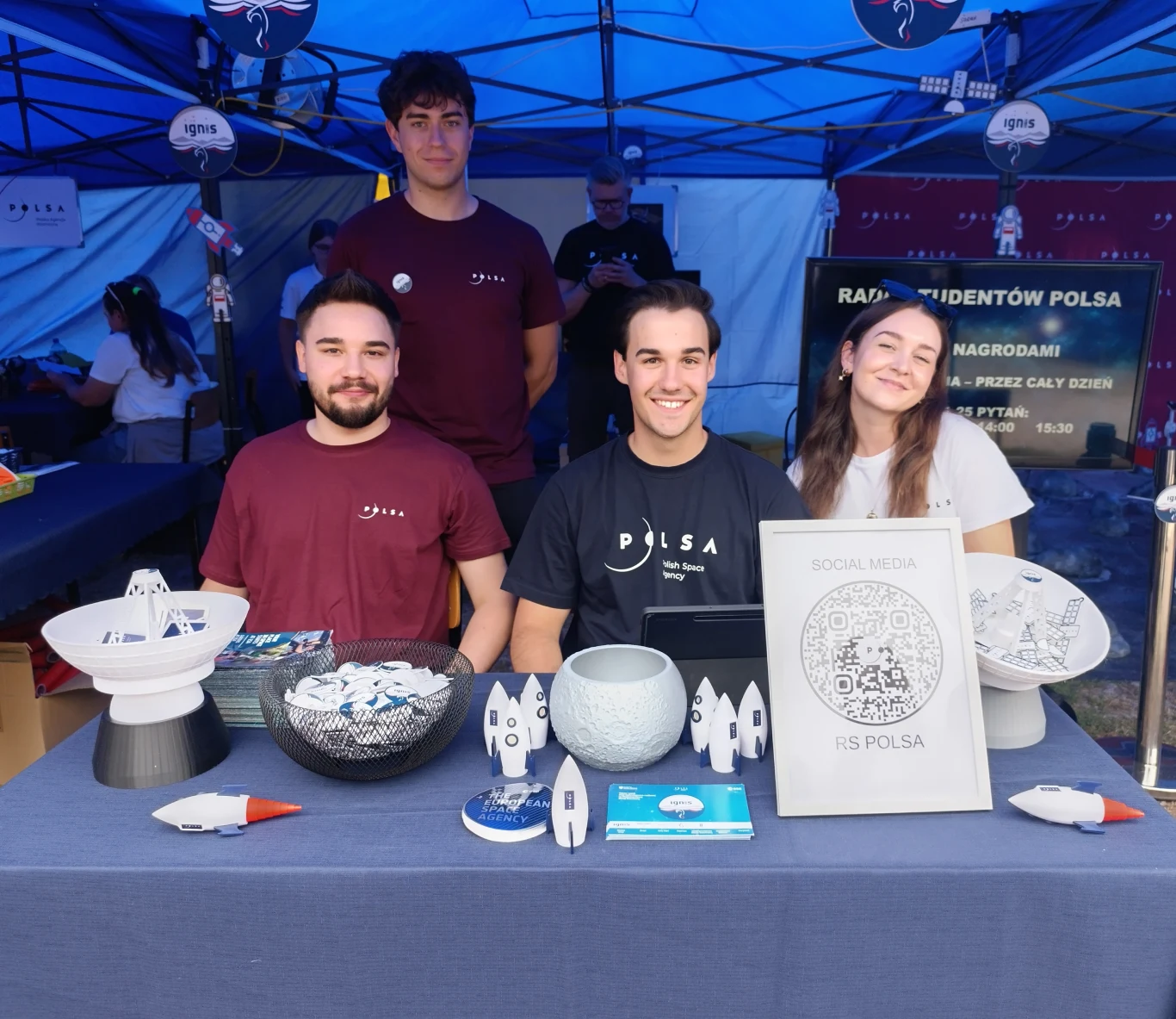 Stanowisko Polskiej Agencji Kosmicznej na ERC 2025 Czterech młodych ludzi uśmiechniętych siedzi i stoi przy stole wystawienniczym pod niebieskim namiotem, na stole przed nimi znajdują się modele rakiet, broszury, miski z przypinkami i tabliczka informacyjna RIS POLSA, w tle widoczne plakaty i dekoracje...