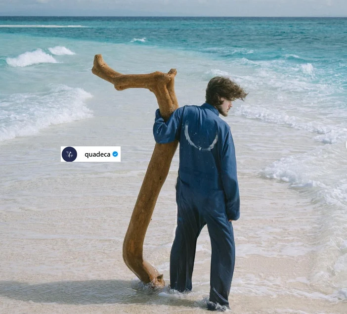 Pod pseudonimem Quadeca kryje się Benjamin Lasky Mężczyzna w niebieskim kombinezonie stoi tyłem na brzegu morza, trzymając duży kawałek drzewa przypominający literę Y, fale delikatnie opływają piasek wokół jego stóp, w tle rozciąga się błękitna woda oceanu.