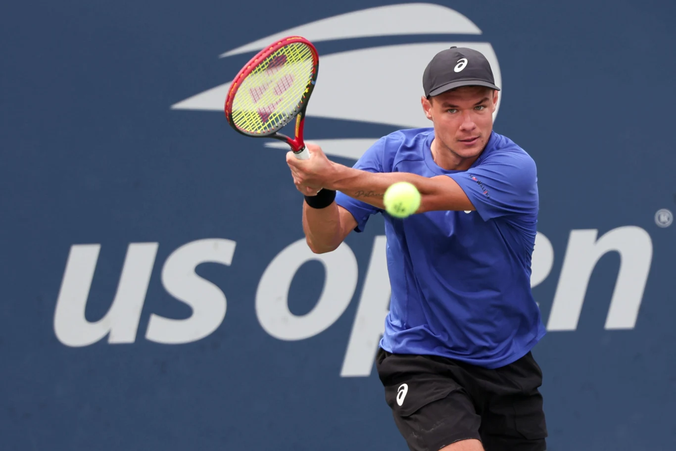 Tenisista w niebieskiej koszulce i czarnej czapce podczas wykonywania uderzenia backhandem na tle logo turnieju US Open.
