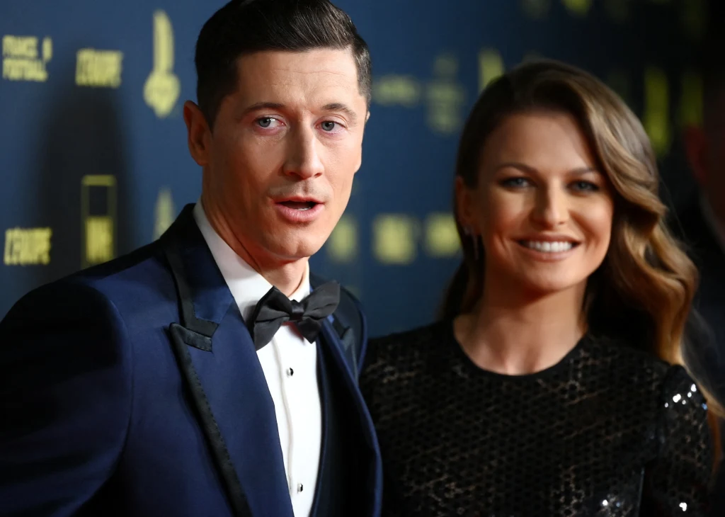 Mężczyzna w eleganckim granatowym smokingu z muchą oraz kobieta w czarnej, błyszczącej sukni stoją razem na tle ścianki z logotypami, oboje uśmiechnięci, w klimacie gali.