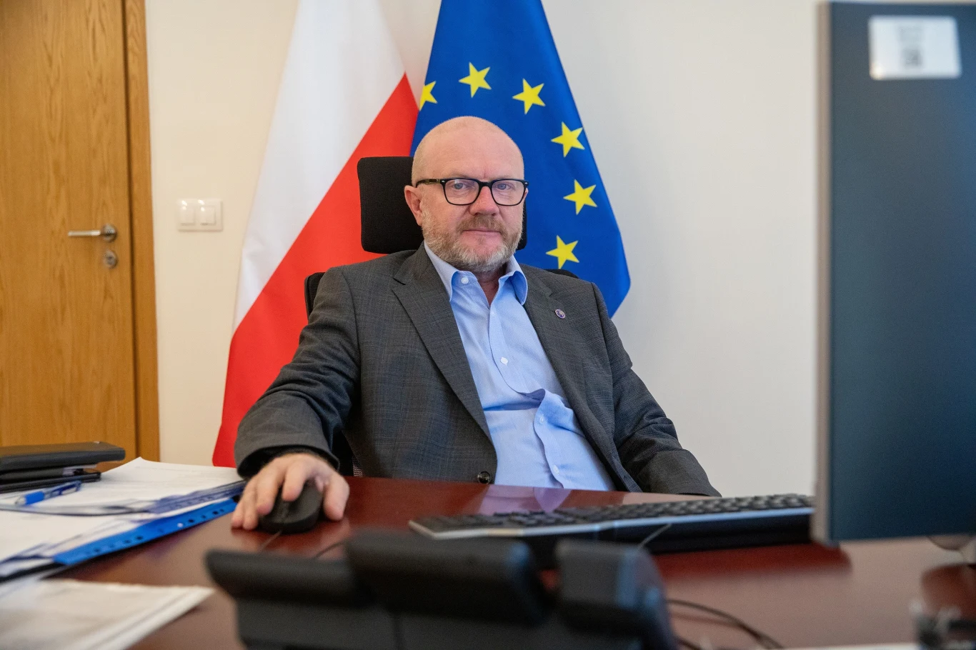 Wiceminister MSWIA Maciej Duszczyk Mężczyzna w garniturze siedzący przy biurku w gabinecie, w tle widoczne flagi Polski i Unii Europejskiej, na biurku komputer i dokumenty, atmosfera formalna i urzędowa.