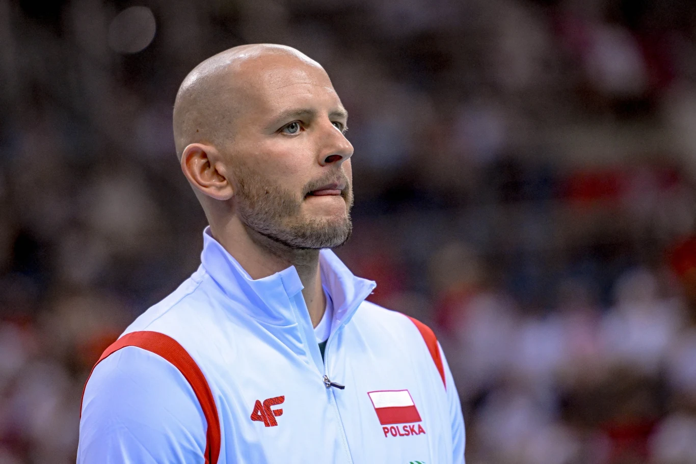 Bartosz Kurek Mężczyzna w białej sportowej kurtce z emblematem Polski oraz logo firmy 4F na tle stadionu lub hali sportowej, skupiony, z poważnym wyrazem twarzy.