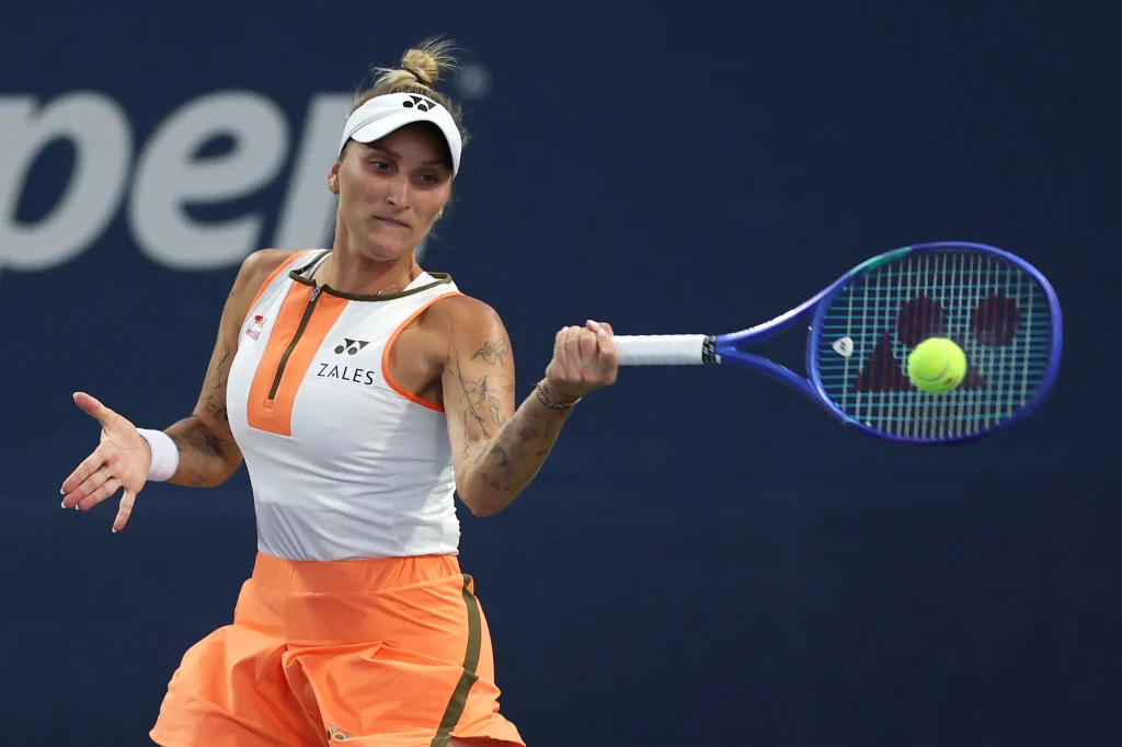 Marketa Vondrousova Tenisistka w trakcie uderzenia piłki podczas meczu, skupiona miną, ubrana w pomarańczowy strój sportowy, w tle niewyraźne logo i ciemnoniebieskie tło kortu.