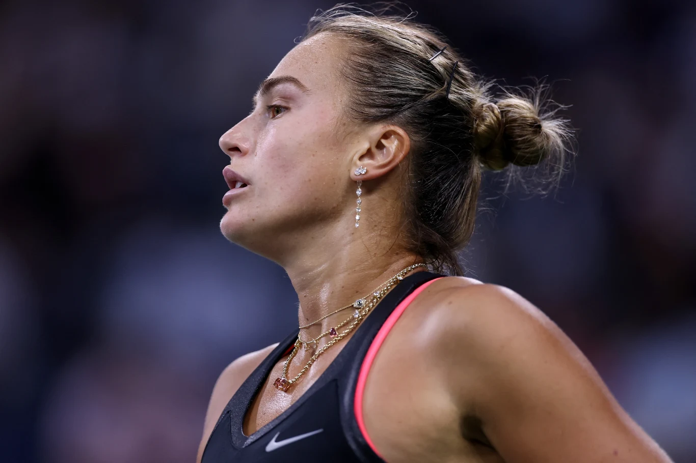 Aryna Sabalenka Kobieta o spiętych włosach ubrana w sportowy strój, z widoczną biżuterią na szyi, znajduje się w sportowym otoczeniu.