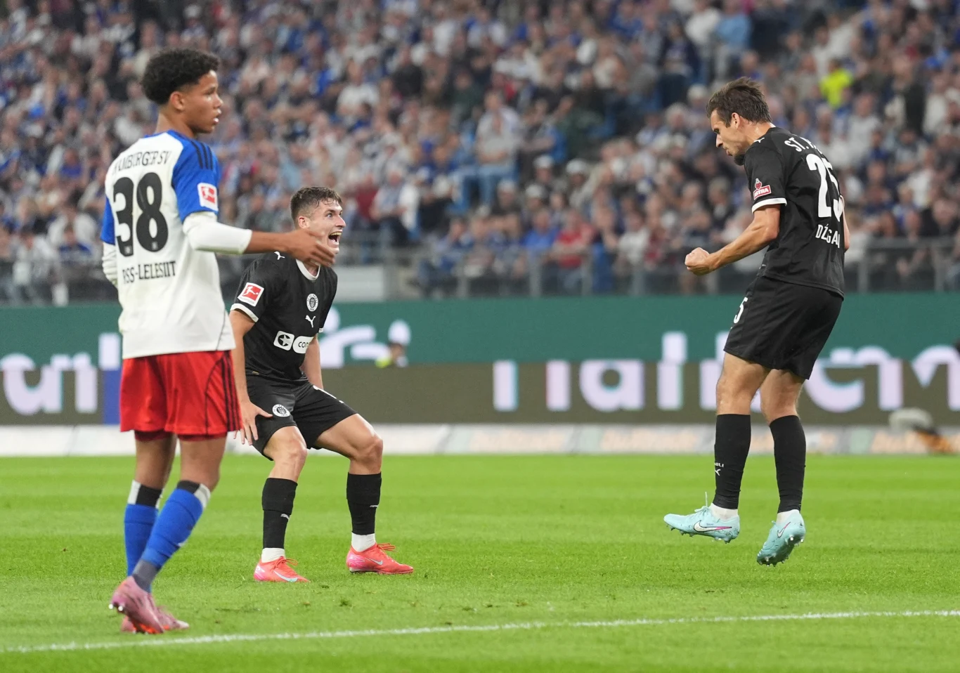 Dwóch piłkarzy w czarnych strojach oraz jeden zawodnik drużyny przeciwnej w biało-czerwono-niebieskim stroju podczas meczu piłkarskiego na stadionie, na tle trybun pełnych kibiców, jeden z graczy unosi się w podskoku, a drugi wyraża emocje.