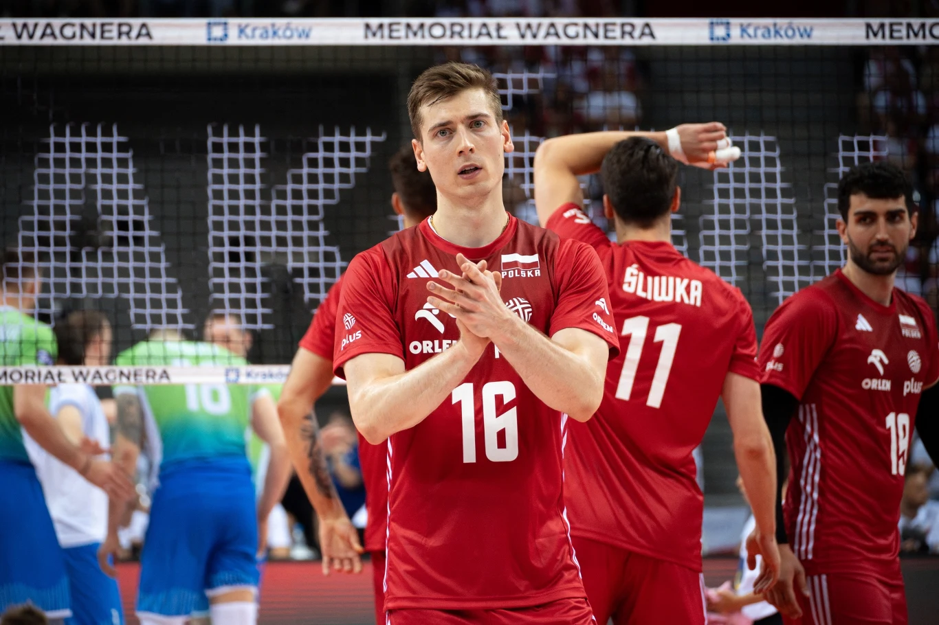 Zawodnik w czerwonym stroju siatkarskim stoi na boisku, klaszcząc w dłonie, w tle siatka oraz pozostali gracze drużyny i przeciwnicy ubrane na niebiesko i zielono, otoczenie hali sportowej z napisem Kraków oraz Memorial Wagnera.