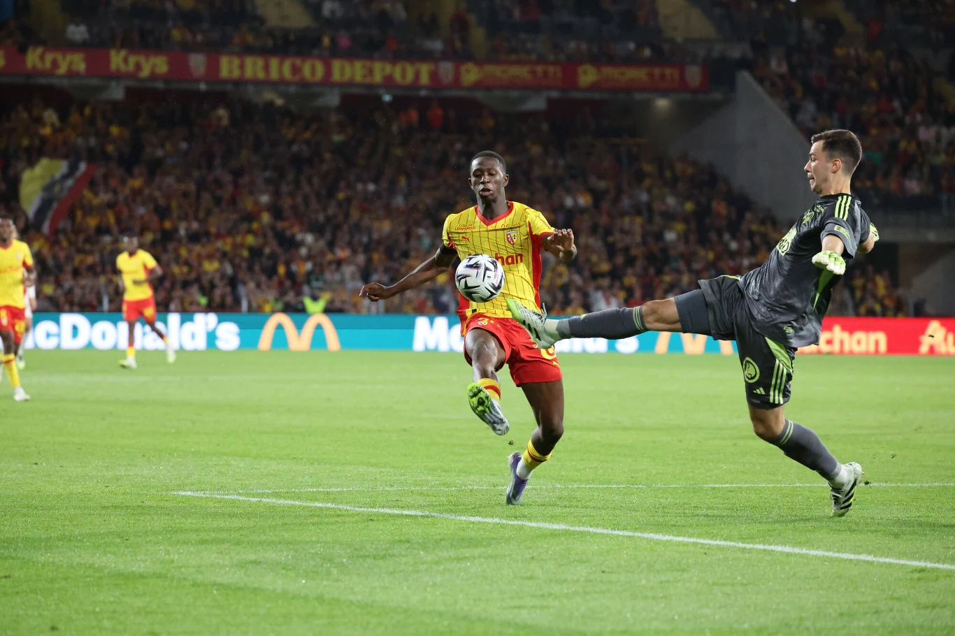 Radosław Majecki w spotkaniu z RC Lens otrzymał czerwoną kartkę. Po jego faulu został podyktowany rzut karny Dwóch piłkarzy walczących o piłkę na murawie stadionu podczas meczu przy pełnych trybunach, zawodnik w żółto-czerwonym stroju i bramkarz w czarnym stroju w dynamicznej akcji, atmosfera wieczornego meczu z widoczną publicznością i reklamami w tle.
