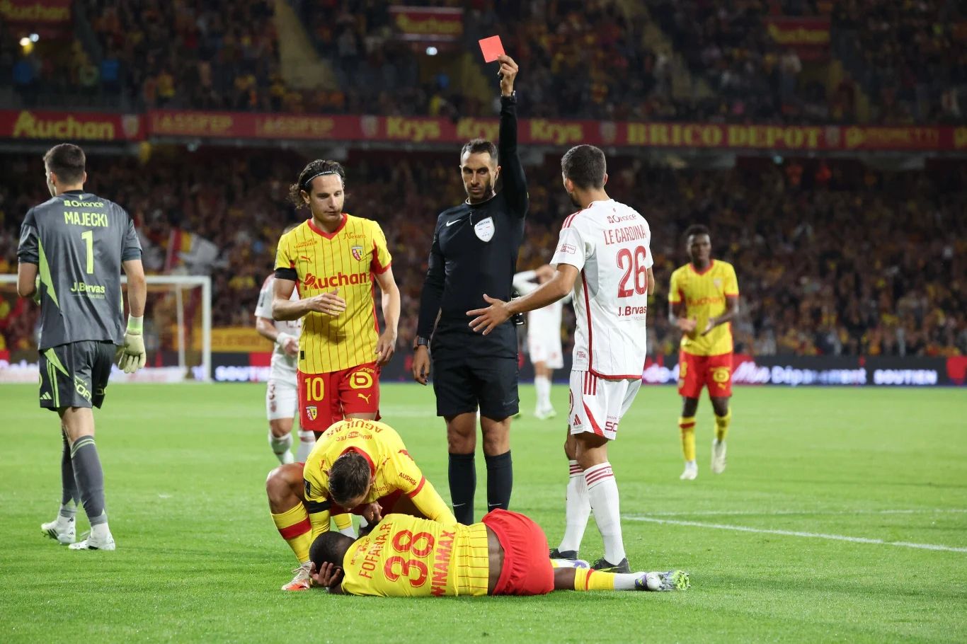Czerwona kartka Radosława Majeckiego w Ligue 1 Sędzia trzymający czerwoną kartkę nad głową, piłkarz drużyny w białym stroju gestykulujący w stronę arbitra, zawodnicy w żółtych strojach, jeden z nich leży na murawie trzymając się za nogę, scena rozgrywa się podczas meczu piłkarskiego przy pełnych tr...