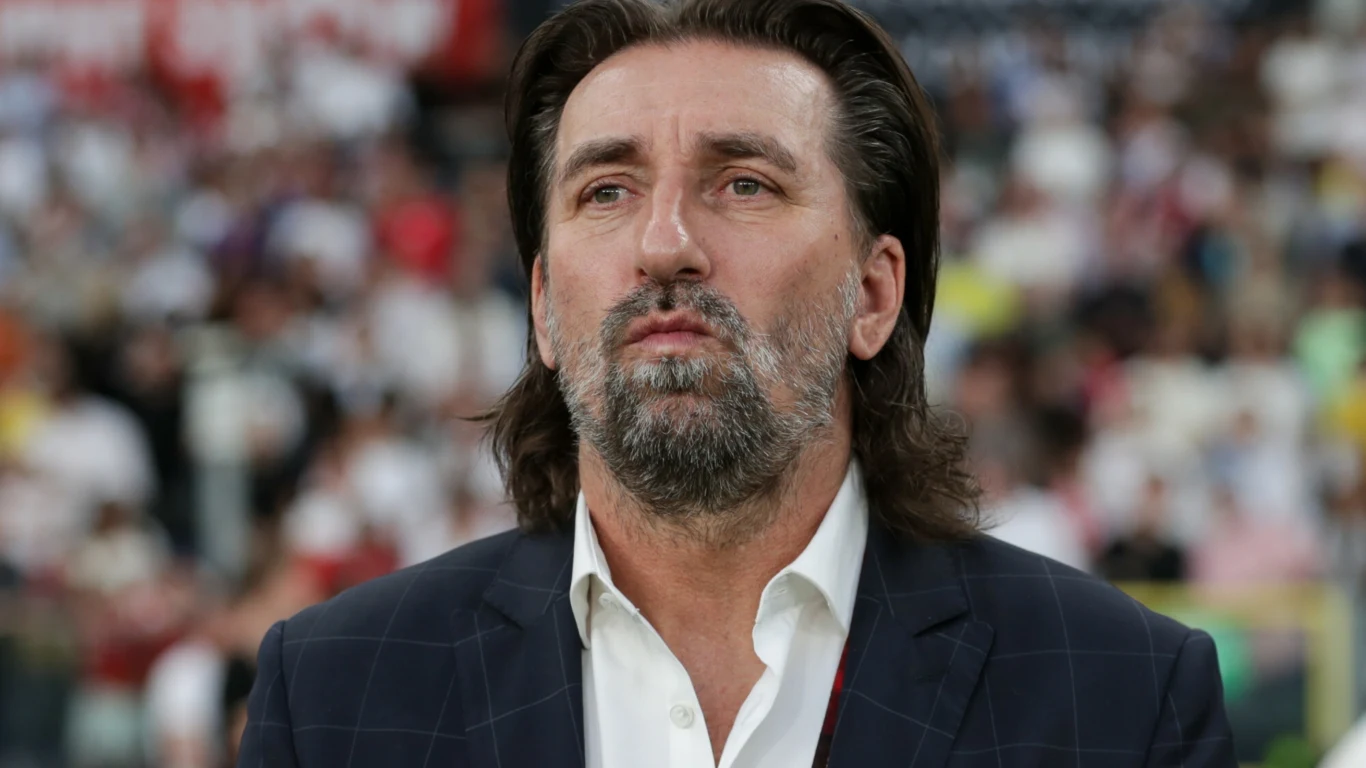 Mężczyzna z dłuższymi ciemnymi włosami i brodą w eleganckiej marynarce, w tle rozmyta publiczność stadionu lub dużego wydarzenia sportowego.