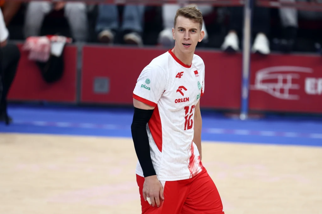 Siatkarz ubrany w biało-czerwony strój sportowy z numerem 16 stoi na boisku podczas meczu halowego, w tle widoczne trybuny oraz elementy logotypu sponsora.