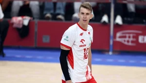 Siatkarz ubrany w biało-czerwony strój sportowy z numerem 16 stoi na boisku podczas meczu halowego, w tle widoczne trybuny oraz elementy logotypu sponsora.