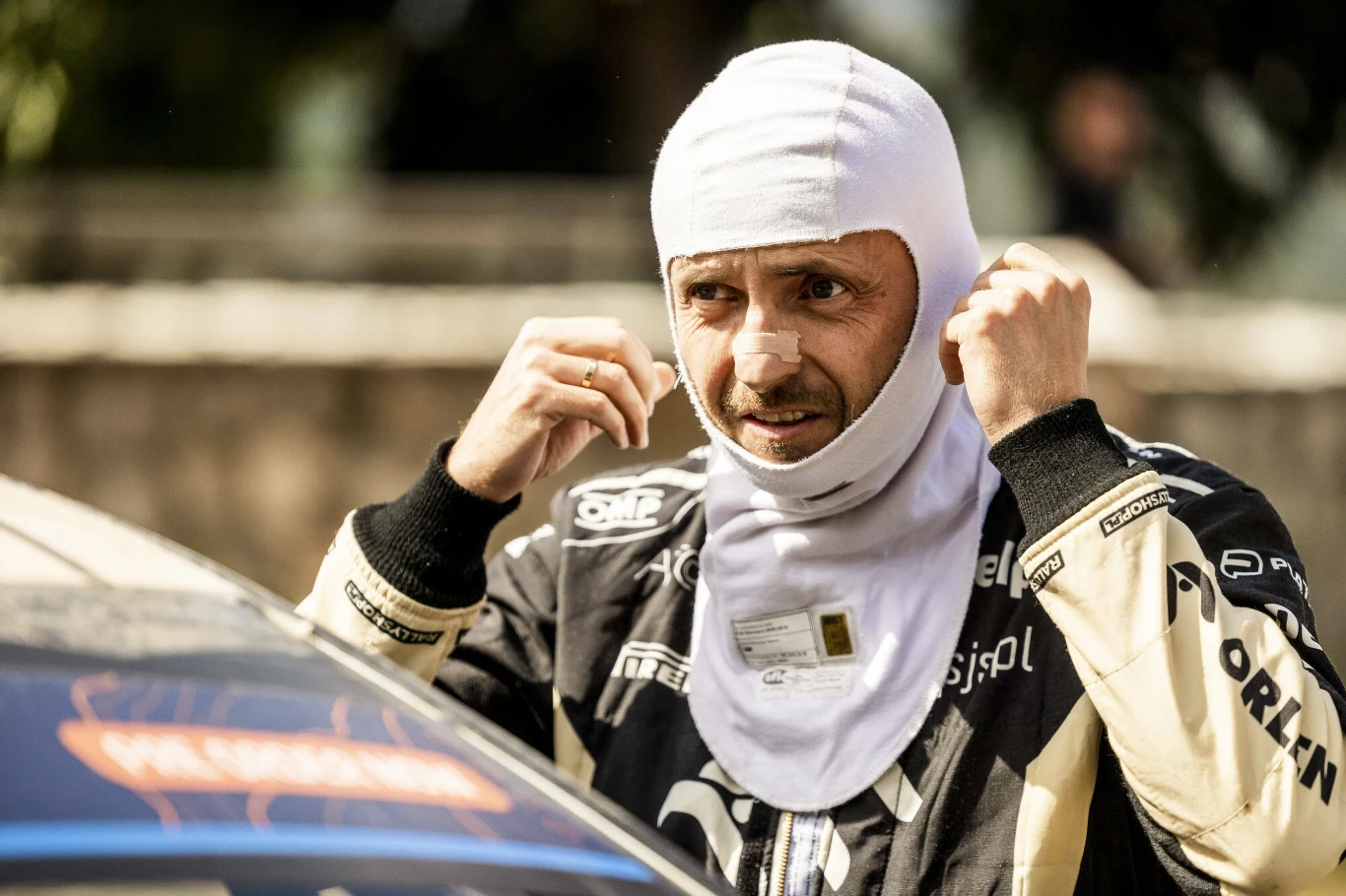 Kierowca rajdowy zakładający balaklawę, stojący obok samochodu wyścigowego, ubrany w kombinezon z widocznymi logotypami sponsorów.