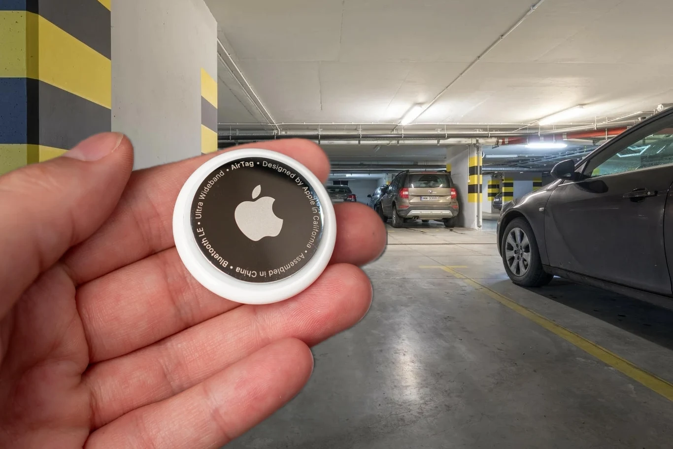 Osoba trzyma lokalizator Apple AirTag w podziemnym parkingu, w tle widoczne są zaparkowane samochody oraz oznaczenia na ścianach i filarach.