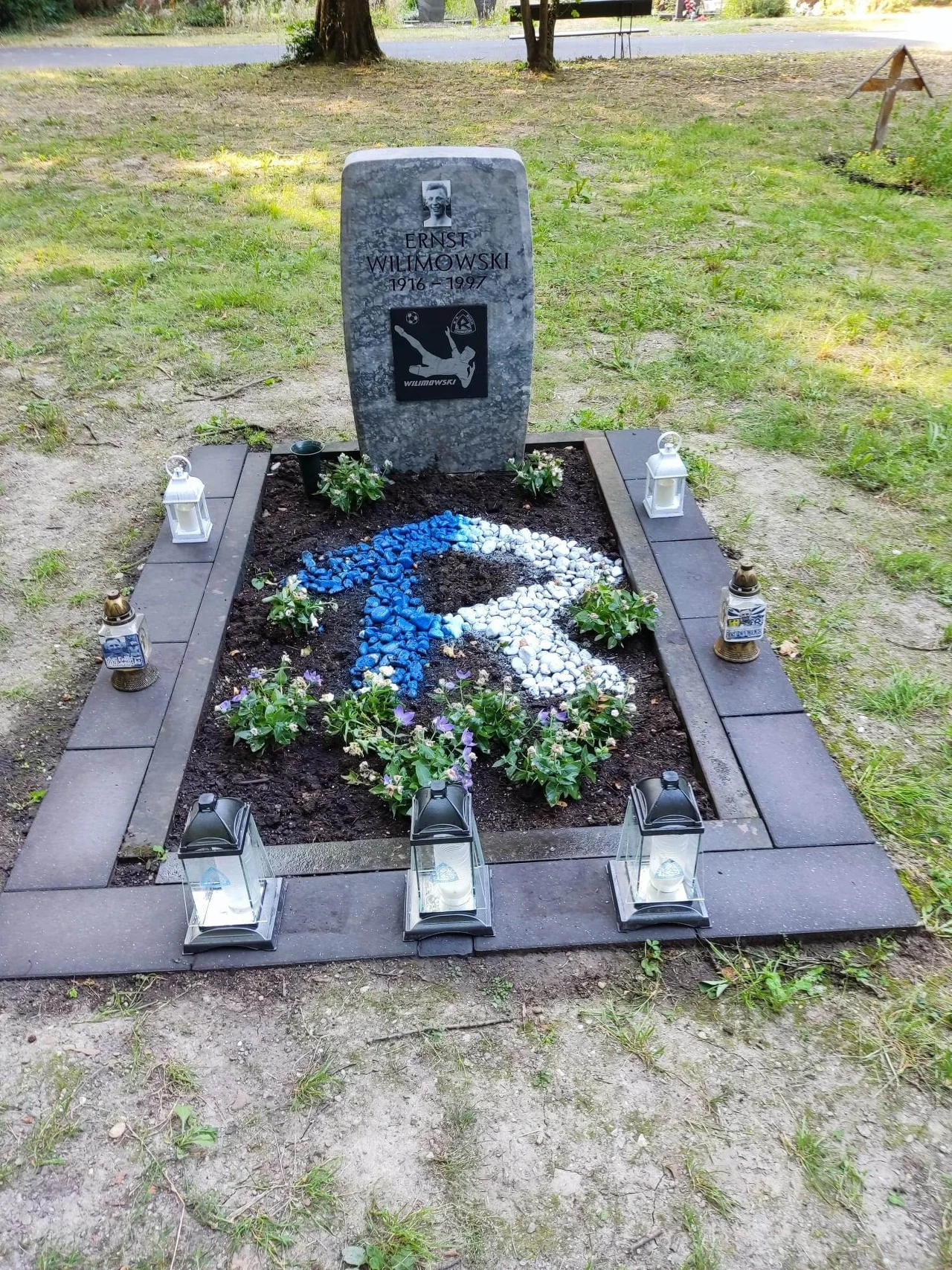 Nagrobek z napisem, zdjęciem oraz ozdobami, otoczony świeczkami i roślinami. Na grobie kolorowa mozaika z kamieni w kształcie symbolu przypominającego orła, wszystko otoczone jasnym obramowaniem.