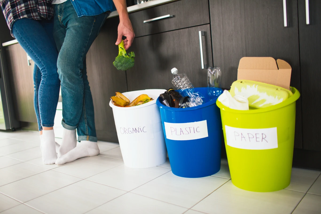 Segregacja odpadów bywa skomplikowana i nieoczywista Trzy kosze do segregacji odpadów kuchennych oznaczone jako organiczne, plastikowe i papierowe wraz z widocznymi odpadami, obok stoją dwie osoby, z których jedna trzyma brokuł nad pojemnikiem.