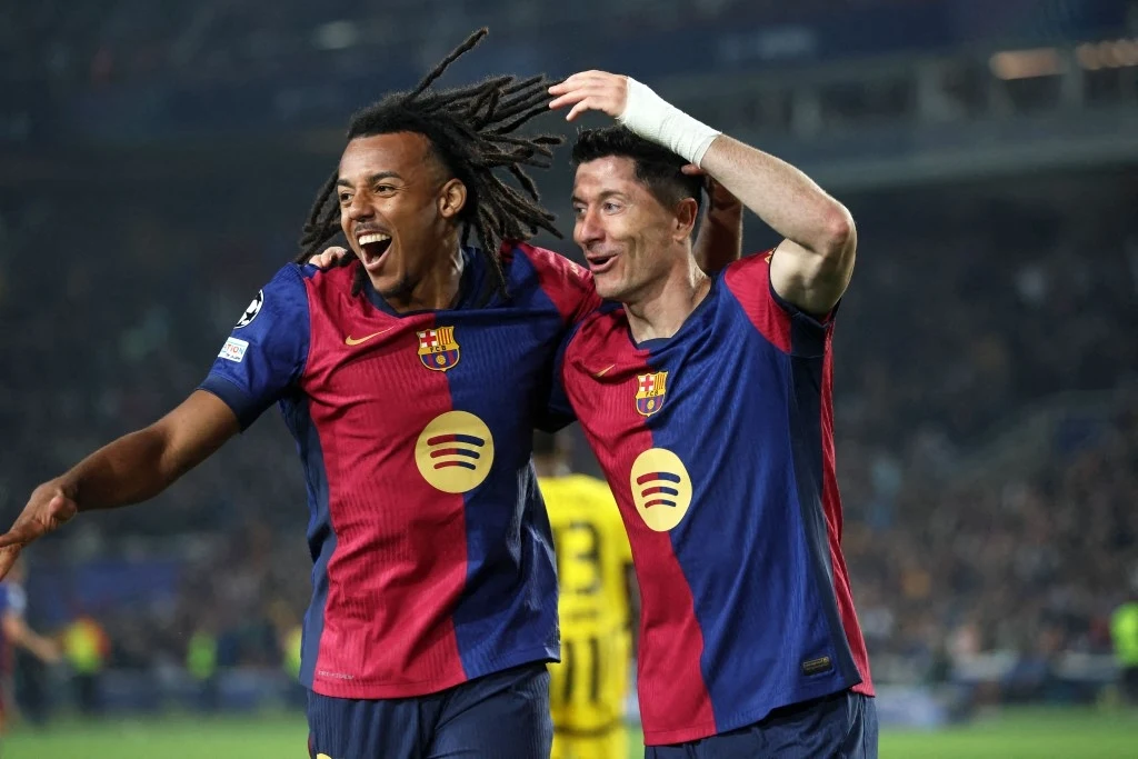 Dwóch piłkarzy FC Barcelony w charakterystycznych bordowo-granatowych koszulkach świętuje zdobycie gola na boisku, jeden z nich z uniesioną ręką, uśmiechnięci i otoczeni kibicami na stadionie