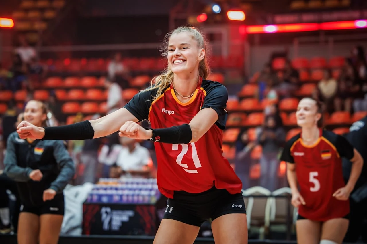 Siatkarki w sportowych strojach wykonujące radosny taniec lub ćwiczenia rozgrzewkowe na hali sportowej, w tle widoczne trybuny oraz oświetlenie sceniczne.