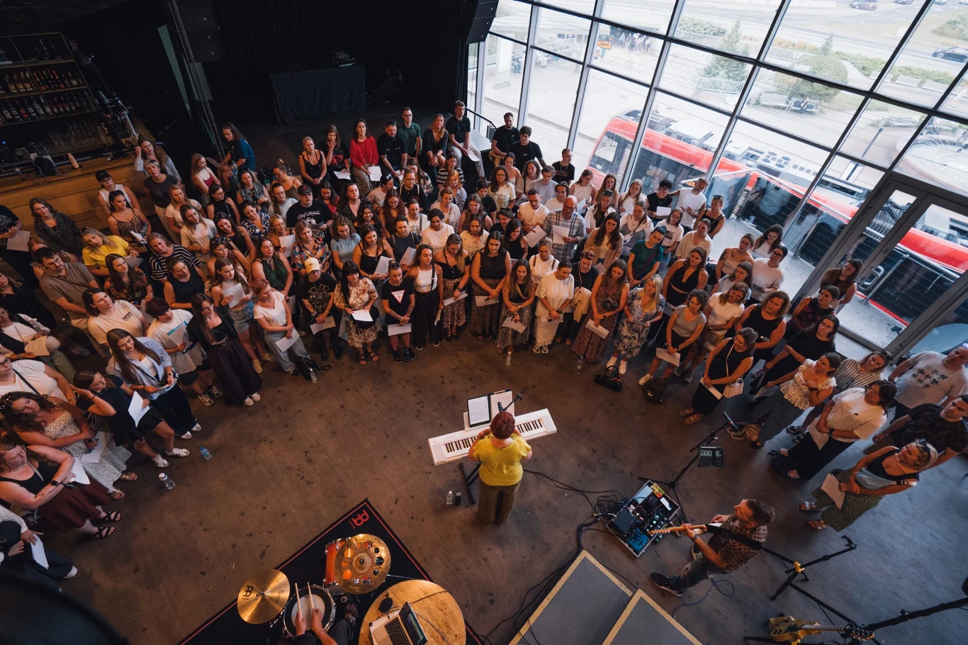 Grupa ludzi zebrana wokół osoby kierującej chórem stojącej przy klawiaturze, po lewej stronie widoczne są instrumenty muzyczne, a całość odbywa się w jasnym pomieszczeniu z dużymi oknami spojrzeniem skierowanym na scenę.