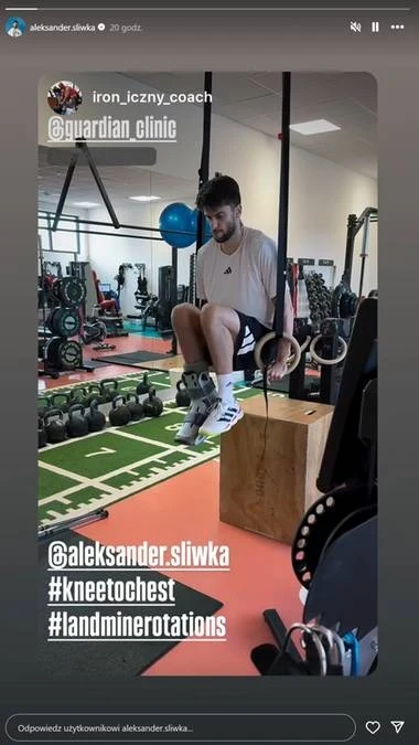 Mężczyzna wykonujący ćwiczenie gimnastyczne na poręczach w profesjonalnie wyposażonej siłowni, otoczony sprzętem sportowym i akcesoriami treningowymi, w tle widoczne lustra oraz maty sportowe.