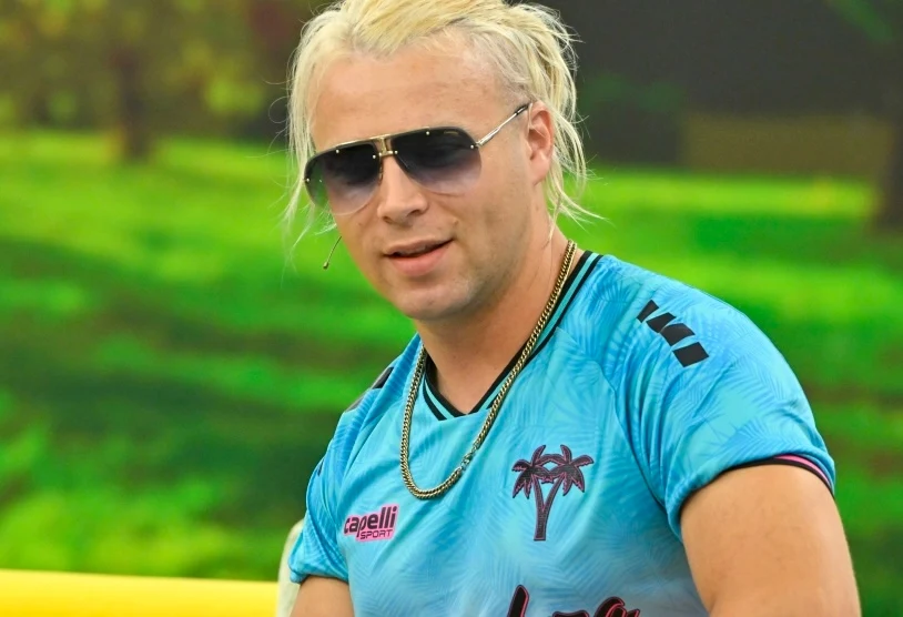 Skolim w "halo tu polsat" opowiedział o swoim hobby Mężczyzna z blond włosami w okularach przeciwsłonecznych, ubrany w sportową niebieską koszulkę z palmami oraz złoty łańcuszek, stoi na tle zielonego, rozmytego krajobrazu.