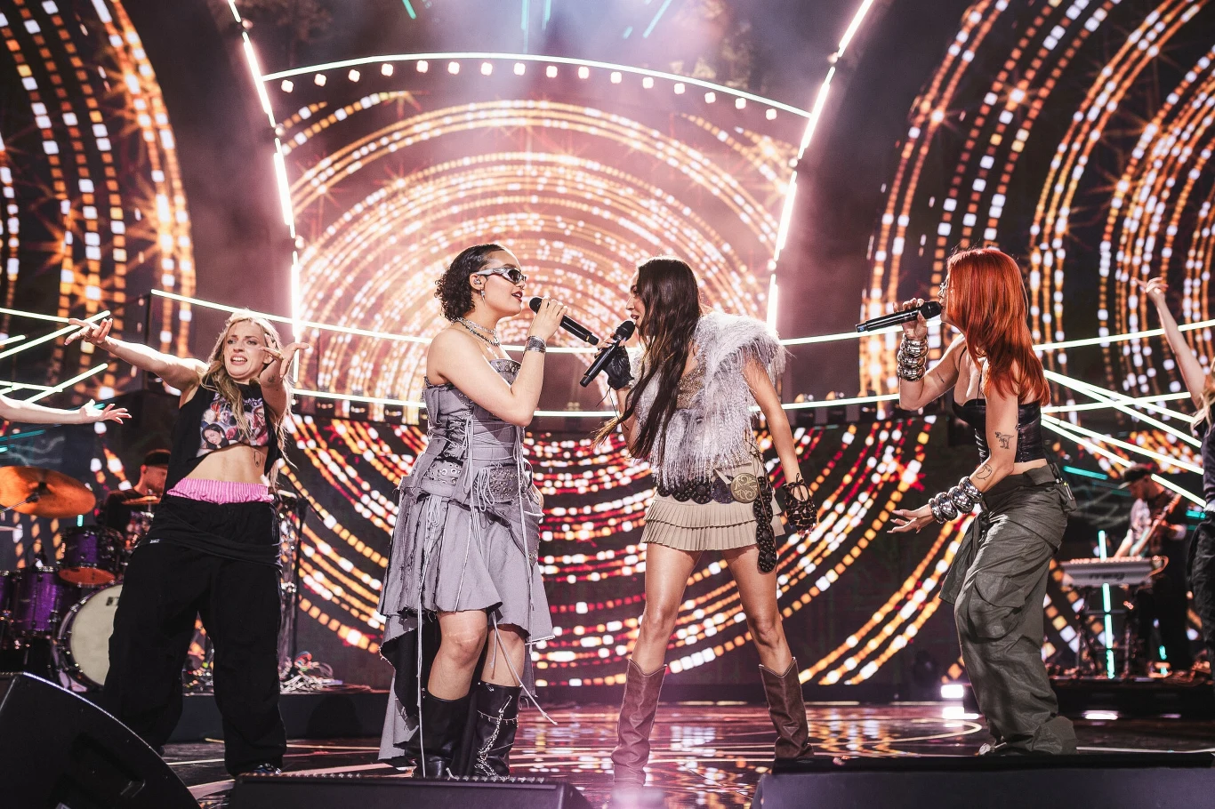 Cztery śpiewające kobiety na scenie w dynamicznych pozach, w otoczeniu wyjątkowych, kolorowych świateł i LED-owych wizualizacji tworzących energetyczną atmosferę koncertu.