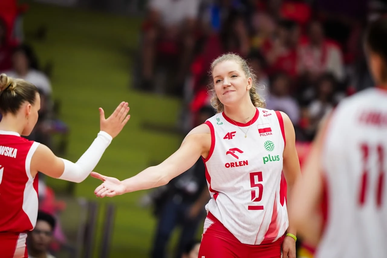 Zawodniczki polskiej reprezentacji siatkówki w trakcie wymiany piątek podczas meczu; w tle rozmyci kibice. Na pierwszym planie widoczna sportsmenka z numerem 5 w biało-czerwonym stroju.