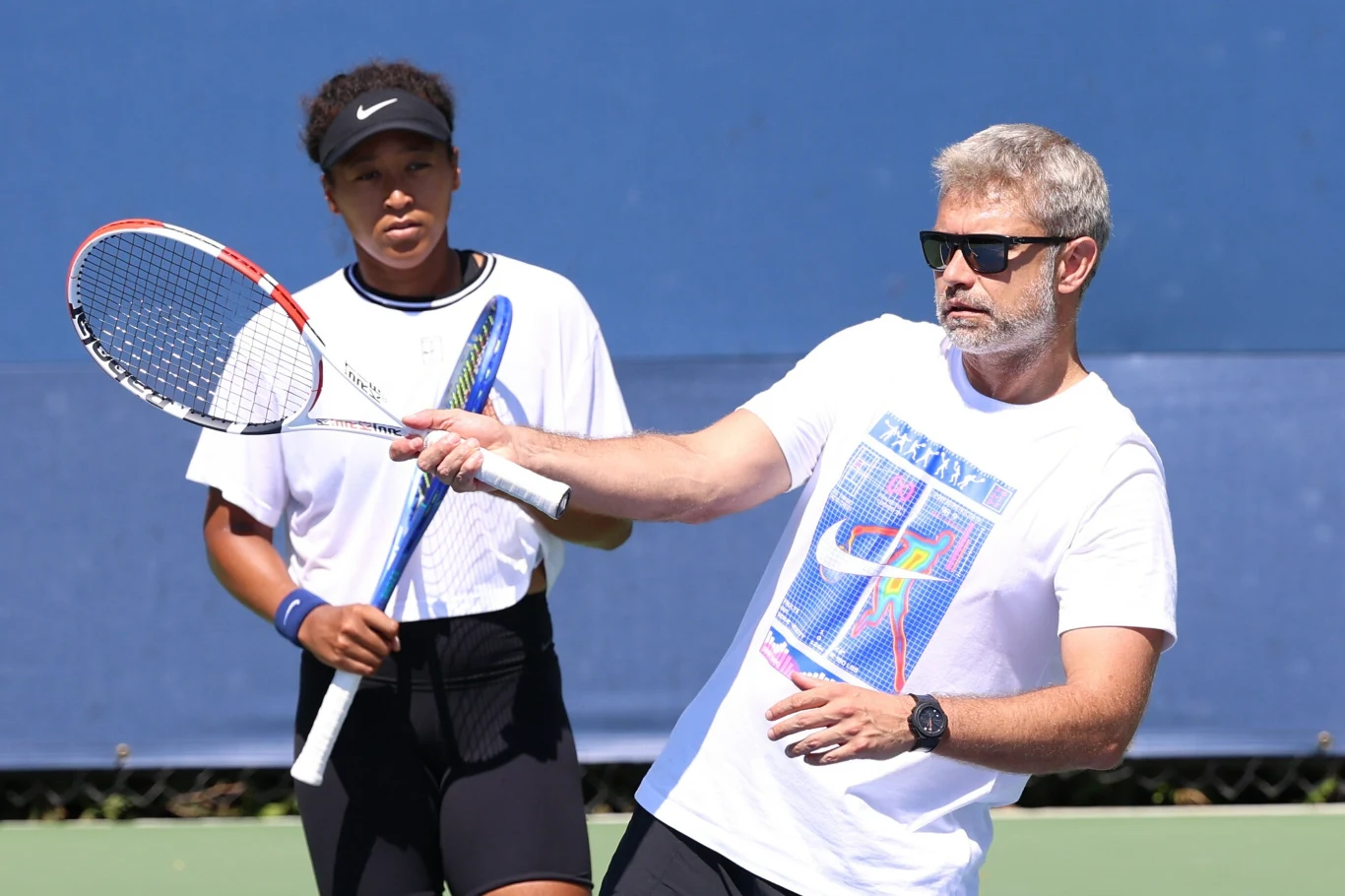 Mężczyzna w okularach przeciwsłonecznych wykonuje ruch rakietą tenisową podczas lekcji na korcie, a obok stoi młoda tenisistka, obserwując jego działanie.