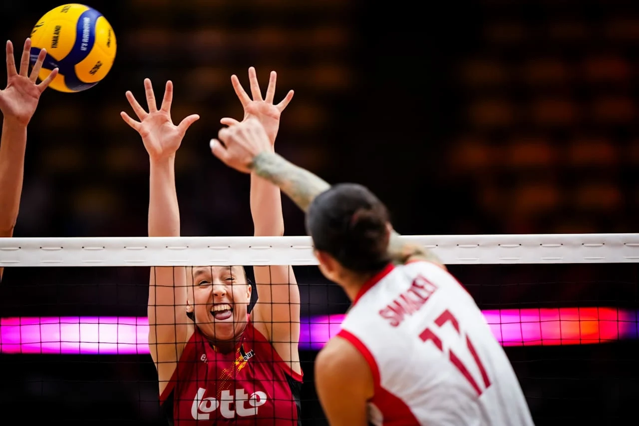 Rywalizacja dwóch zawodniczek podczas meczu siatkówki tuż przy siatce, jedna z nich blokuje atak przeciwniczki, widoczne dynamiczne gesty rąk oraz piłka tuż nad siatką.