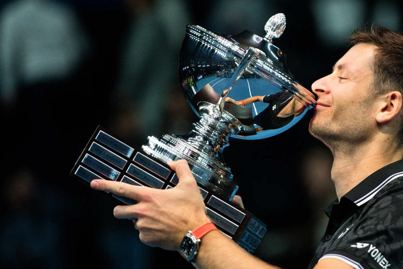 Hubert Hurkacz z pucharem za triumf w Marsylii w 2023 roku Sportowiec w czarnej koszulce obejmuje i całuje dużego, srebrnego pucharu z licznymi tabliczkami, wyrażając radość ze zwycięstwa.