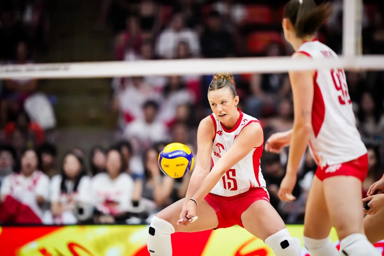Zawodniczka drużyny siatkarskiej ubrana w biało-czerwony strój wykonuje odbiór piłki podczas meczu, w tle wyróżniają się kibice i rozmyte trybuny.