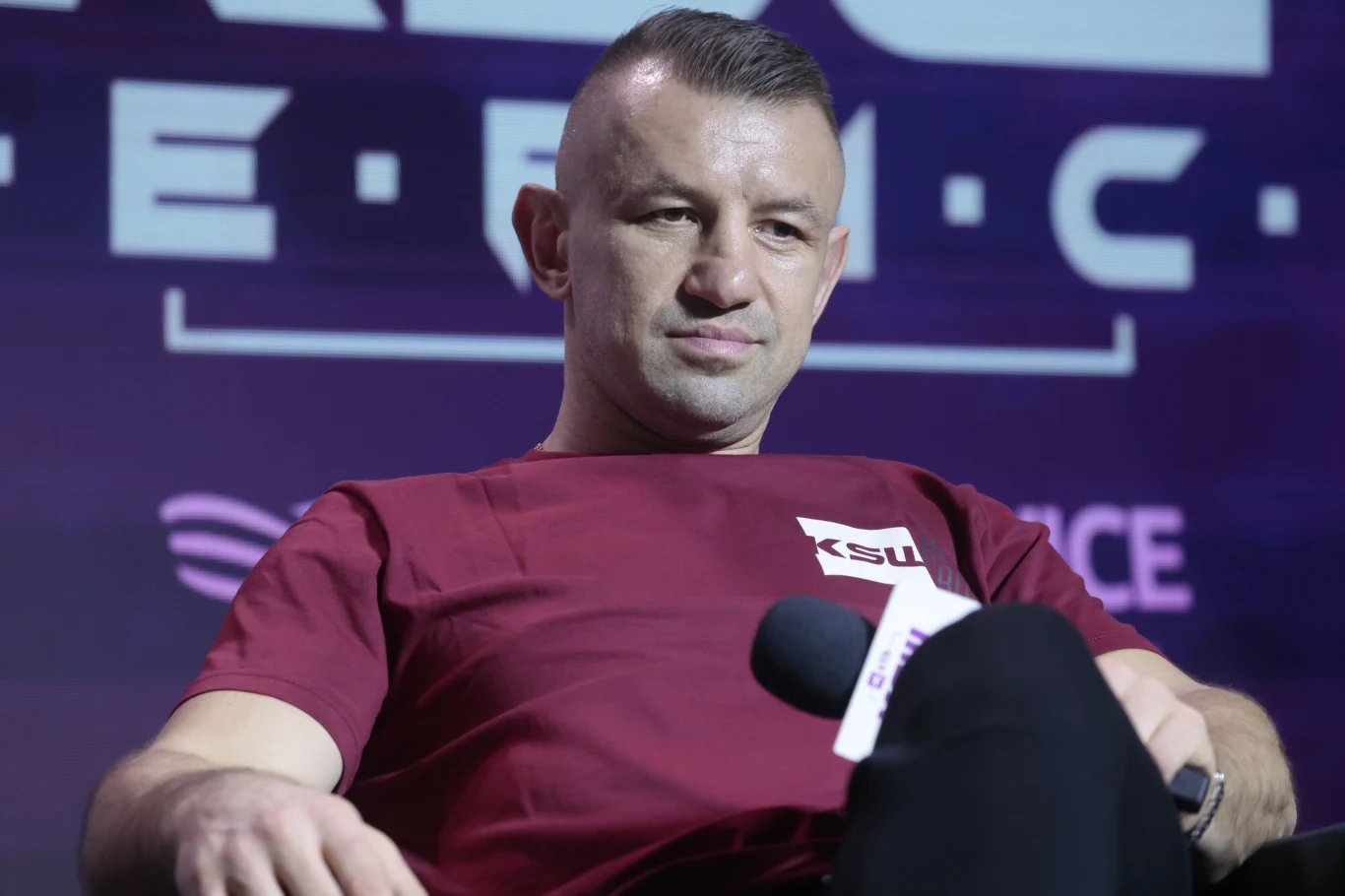 Mężczyzna w bordowej koszulce z napisem KSW siedzi na tle z napisem, trzyma mikrofon, tło ma elementy graficzne oraz litery.