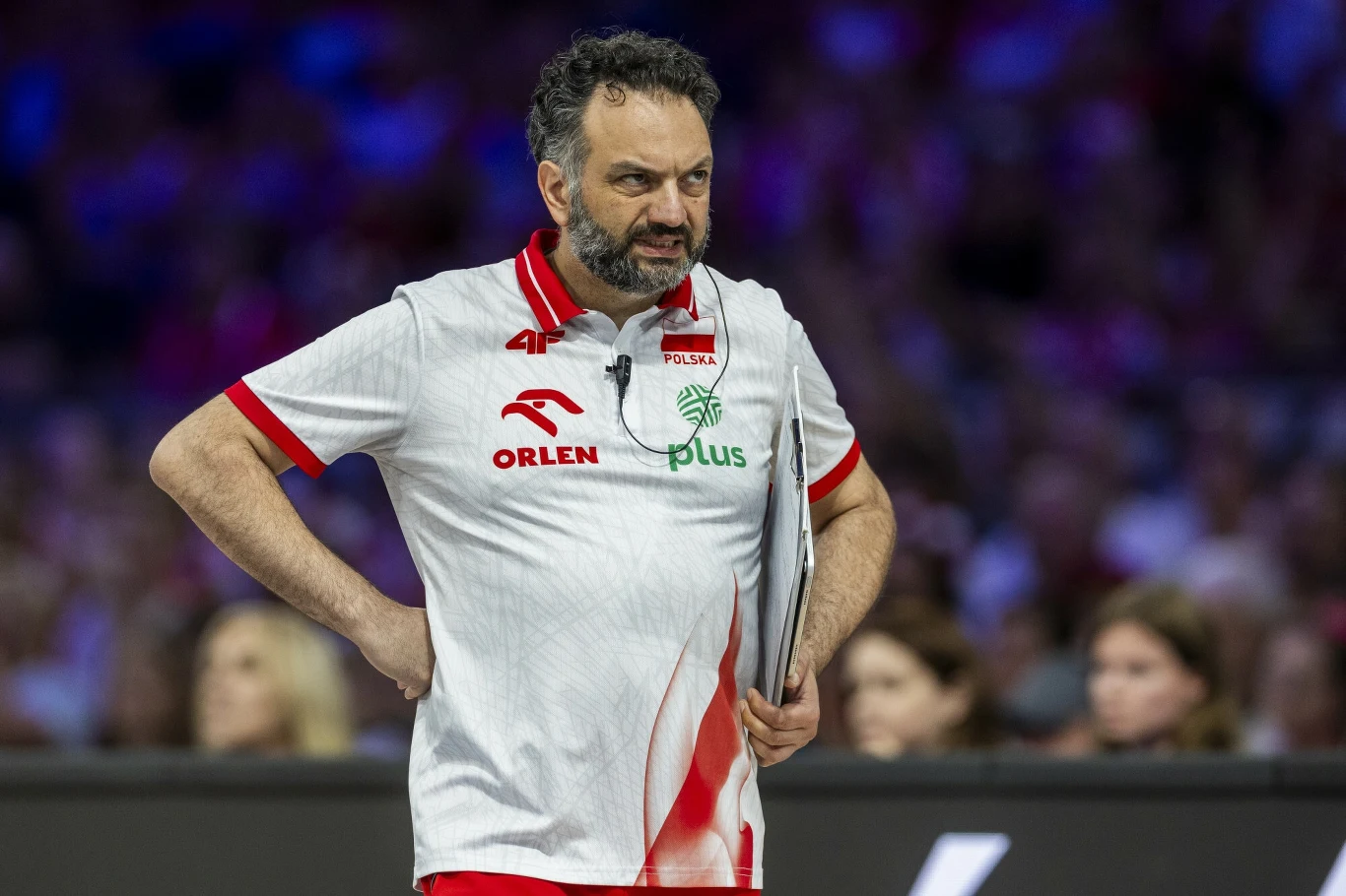 Stefano Lavarini Trener polskiej reprezentacji siatkówki ubrany w biało-czerwoną koszulkę z logotypami sponsorów trzymający dokument lub tablet, stojący na tle rozmazanego tłumu kibiców.