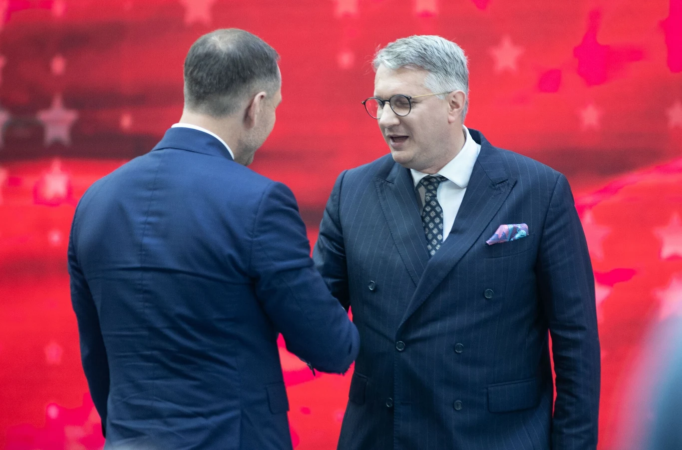 Dwóch mężczyzn w garniturach ściska sobie dłonie na tle czerwonej ściany z jasnymi rozmytymi wzorami, obaj wyglądają na zaangażowanych w rozmowę, jeden z nich ma siwe włosy i okulary.