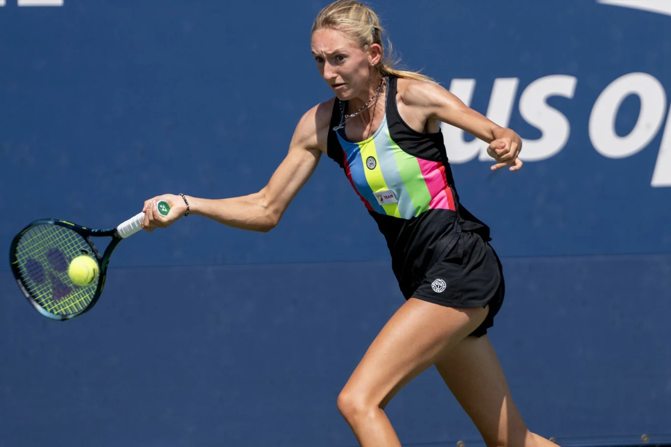 Tenisistka w sportowym stroju wykonuje uderzenie rakietą podczas meczu na niebieskim korcie, w tle widoczne fragmenty napisu US Open.