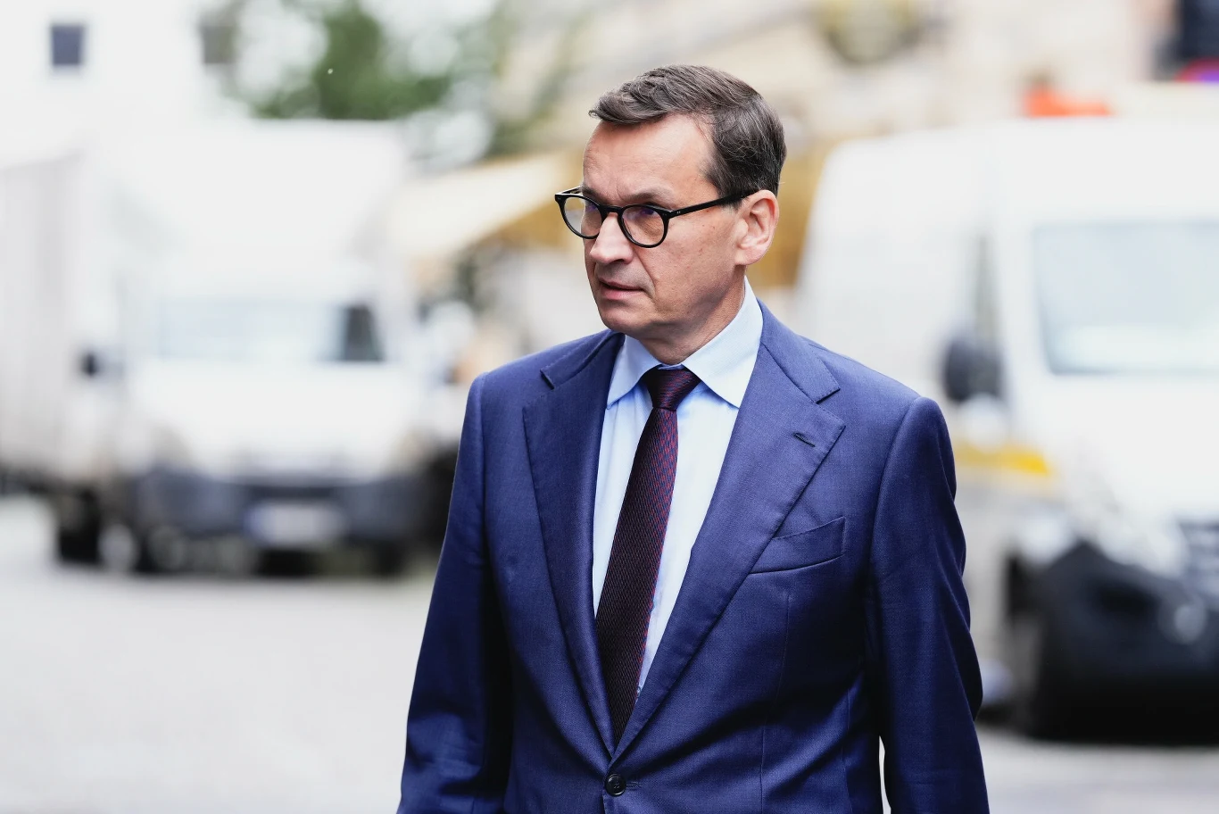 Mężczyzna w garniturze i okularach stoi na ulicy miejskiej, w tle rozmyte samochody dostawcze i fragmenty budynków.